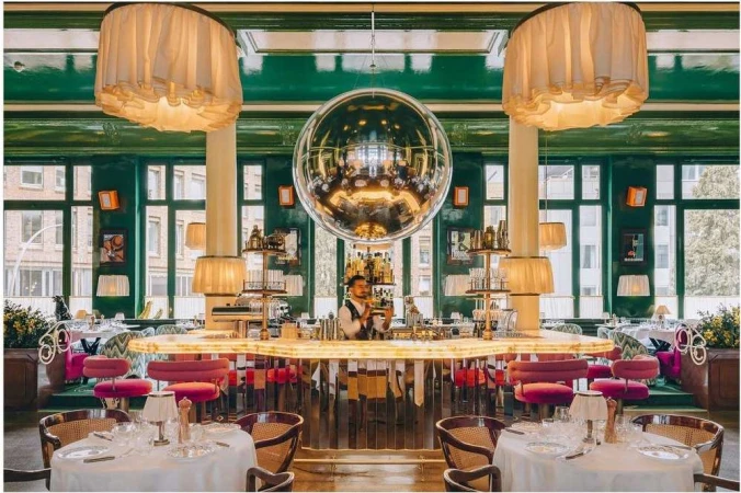 Upscale restaurant interior featuring a mirrored disco ball, seated tables with white linens, and a bartender behind a lighted bar.