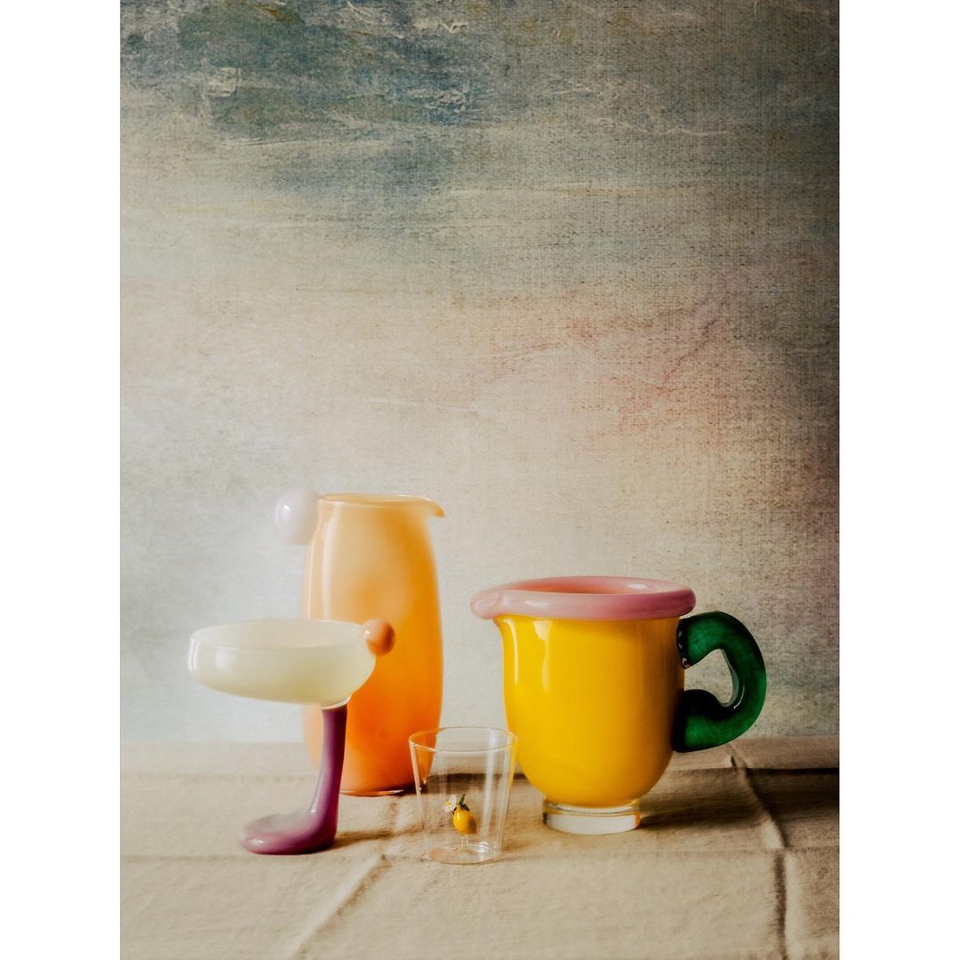 Still life photograph of glass vases and cup in yellow, orange, green, purple, and cream against a painterly backdrop. Food & Beverage.