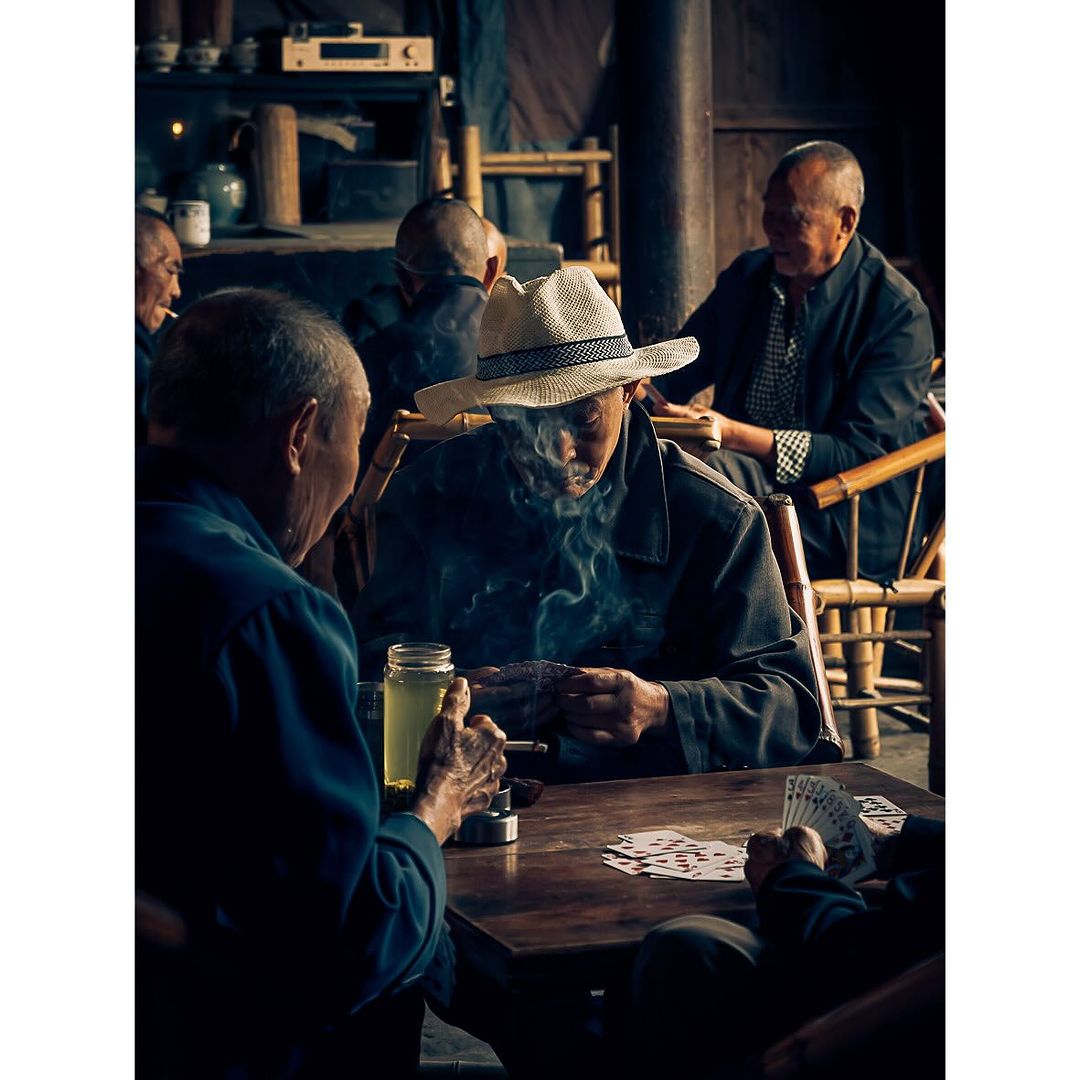 Elderly men play cards and smoke in a dimly lit, rustic indoor setting, with a hat and cigarette smoke as the focal points.
