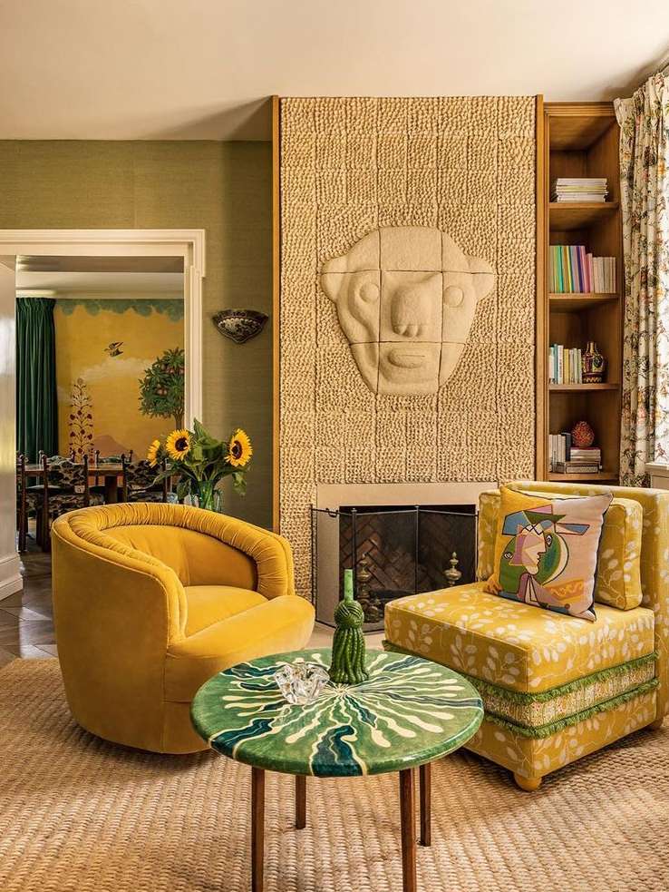 Living room interior with yellow armchairs, a green table, and a fireplace featuring a carved face. Bookshelves and patterned walls complete the design.