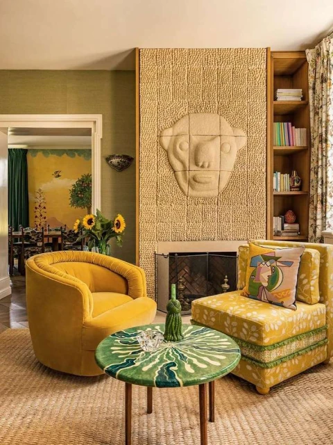 Living room interior with yellow armchairs, a green table, and a fireplace featuring a carved face. Bookshelves and patterned walls complete the design.