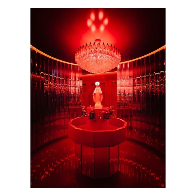 Red-lit interior with sculpture of Virgin Mary on a red octagonal sink, chandelier overhead, and curved, mirrored walls.