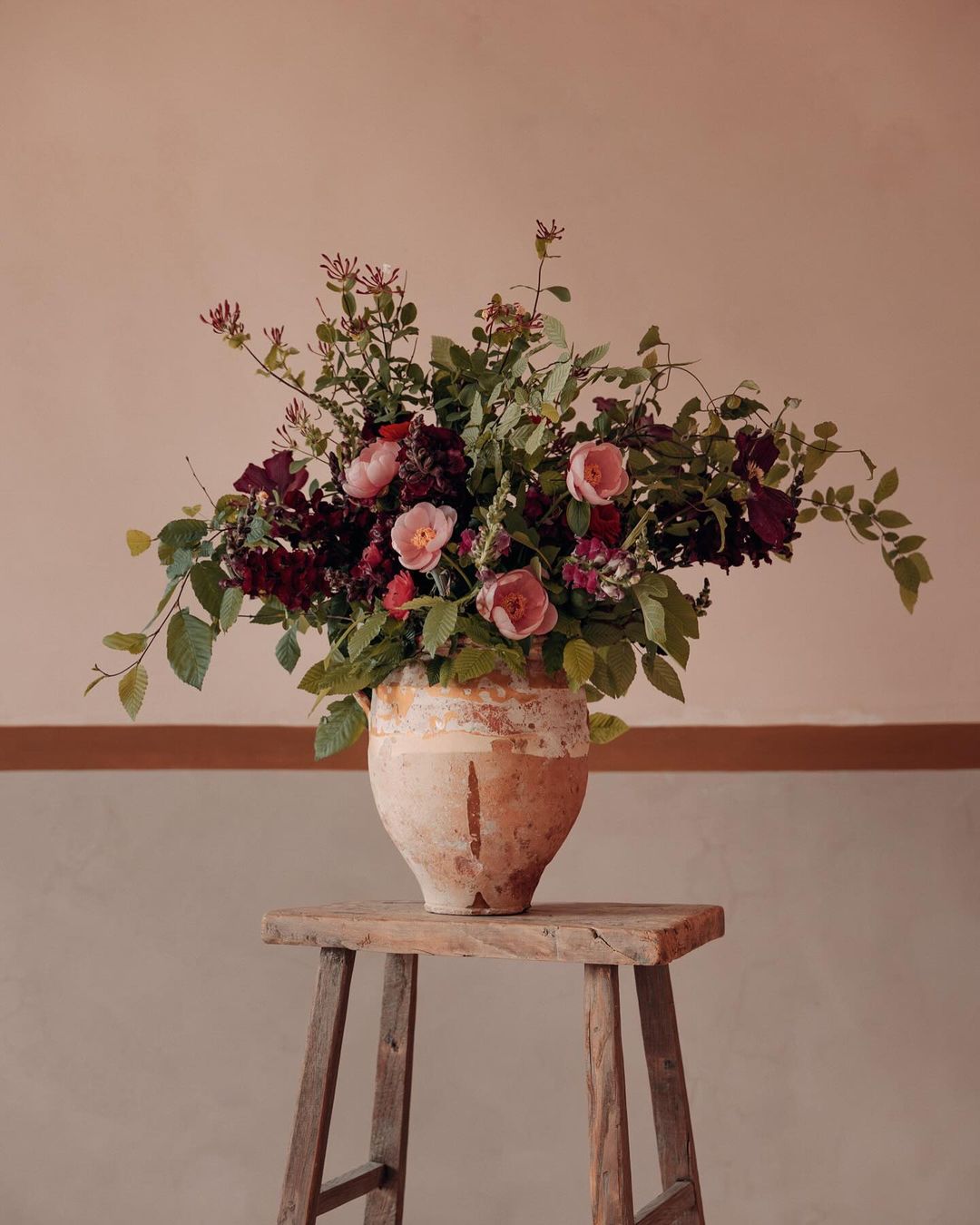 Floral arrangement featuring pink and red roses and green leaves in a weathered vase on a wooden stool against a neutral wall.
