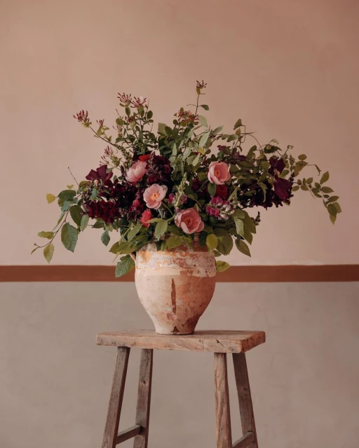 Floral arrangement featuring pink and red roses and green leaves in a weathered vase on a wooden stool against a neutral wall.