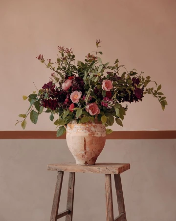 Floral arrangement featuring pink and red roses and green leaves in a weathered vase on a wooden stool against a neutral wall.