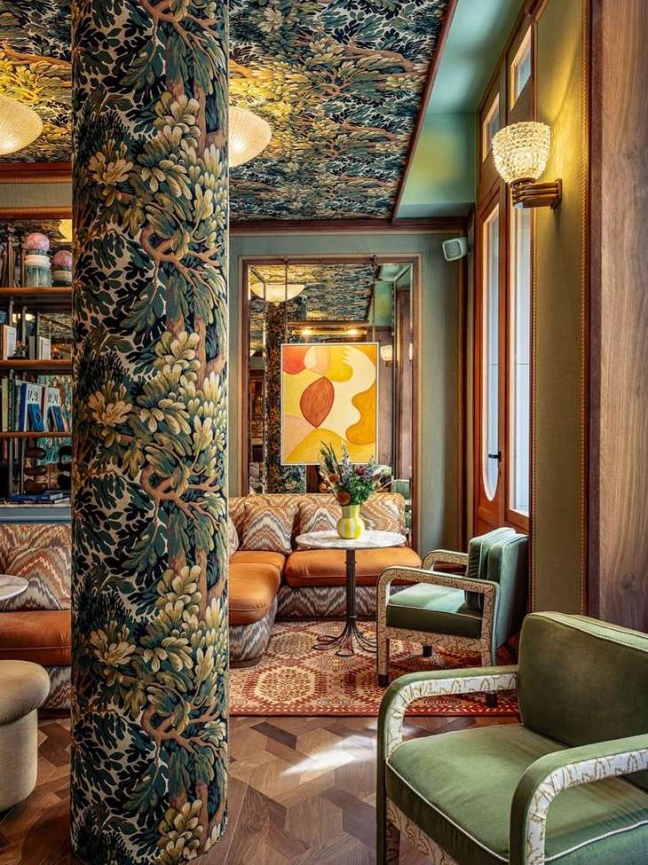 Ornate vintage-style interior of a seating area with a patterned pillar, floral ceiling, an orange couch, and green upholstered armchairs.