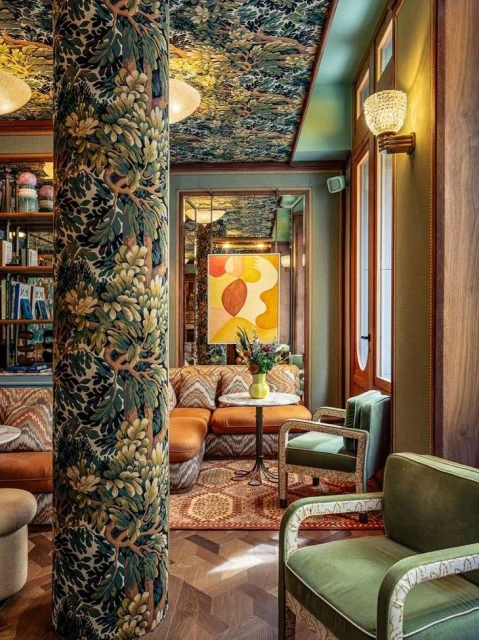 Ornate vintage-style interior of a seating area with a patterned pillar, floral ceiling, an orange couch, and green upholstered armchairs.