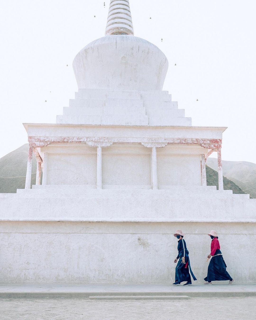 Two people in traditional dress walk past a large white stupa in a serene landscape, featuring overcast lighting and muted tones.