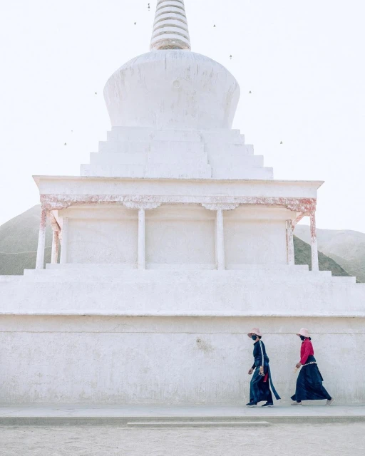 Two people in traditional dress walk past a large white stupa in a serene landscape, featuring overcast lighting and muted tones.