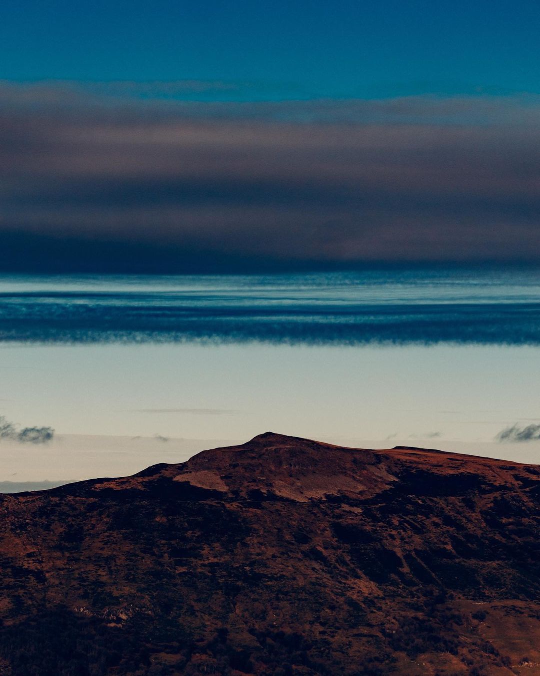 An aerial view showcases a brown mountain under a layered sky of deep blues and whites with a teal-colored horizon providing contrast.