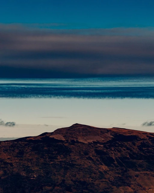 An aerial view showcases a brown mountain under a layered sky of deep blues and whites with a teal-colored horizon providing contrast.