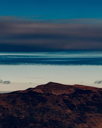 An aerial view showcases a brown mountain under a layered sky of deep blues and whites with a teal-colored horizon providing contrast.