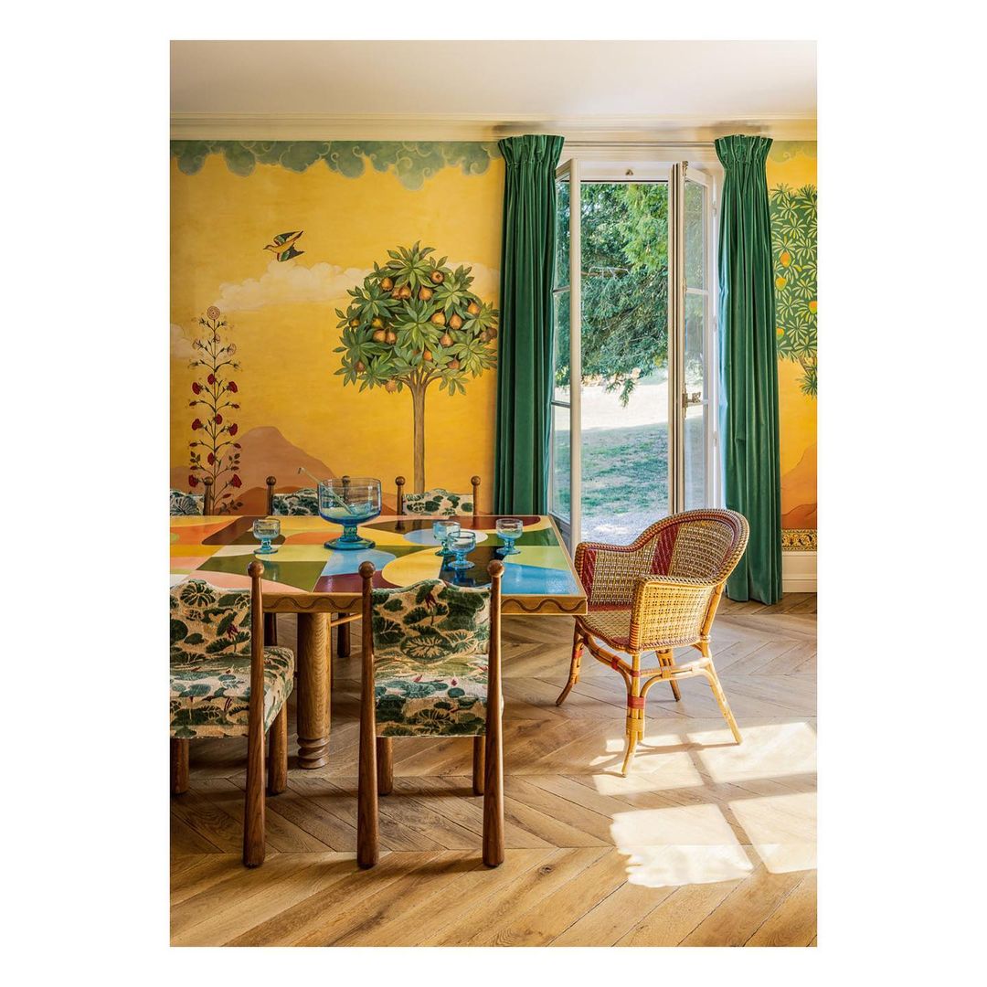 Interior of dining room with colorful table and floral chairs, light wood floor and yellow wallpaper. Light streams in through open door.