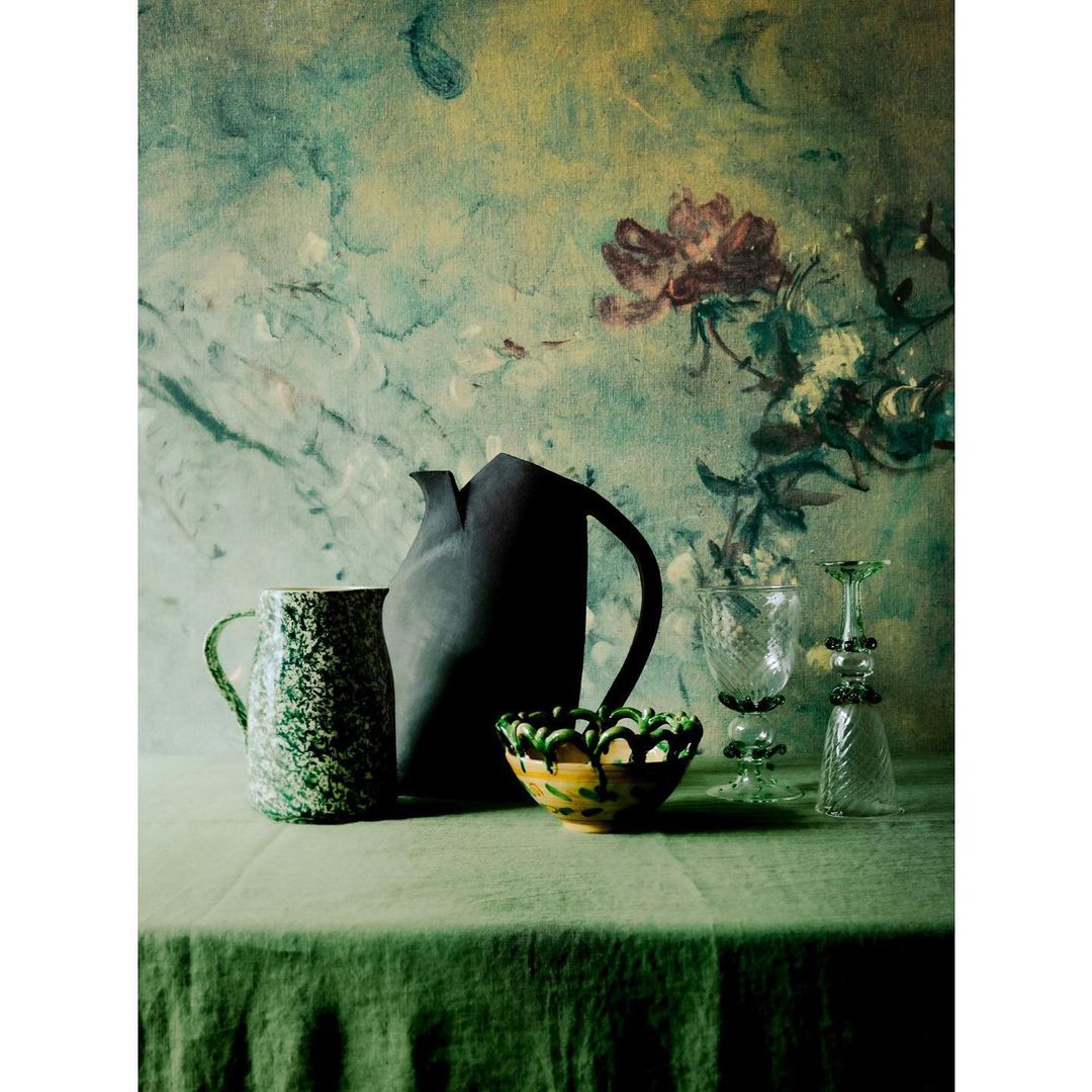 Still life photograph featuring an assortment of pitchers, glasses, and bowls on a green linen tablecloth against an artistic backdrop.