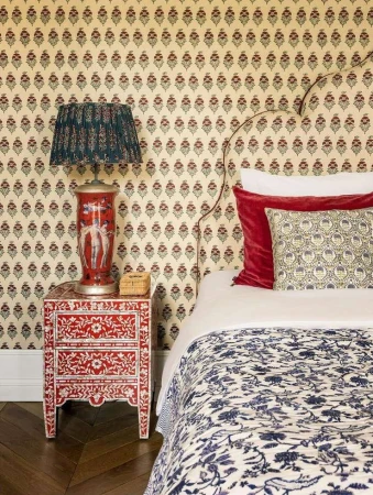 Bedroom interior showcasing a patterned wallpaper, a red bedside table with drawers, a lamp, pillows and a bed with patterned cover.