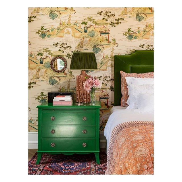 Interior with a green nightstand topped with a lamp, books, and flowers, set against patterned wallpaper and a bed with a green headboard.