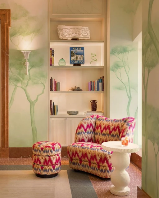 Interior space features a patterned armchair and ottoman, a small round table with a drink, and a bookshelf against a wall with painted trees.