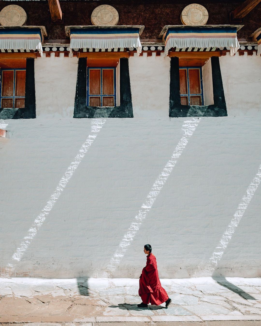 A lone person in red robes walks along a stone path next to a white wall with symmetrical window openings, bathed in sunlight.