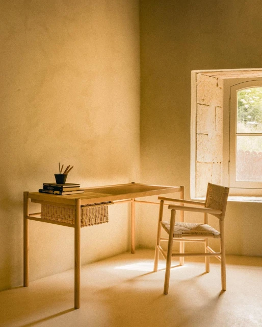Interior shot of a light wood desk with books and pens next to a woven chair near a window, warm color palette, simple, cozy, and minimalist.