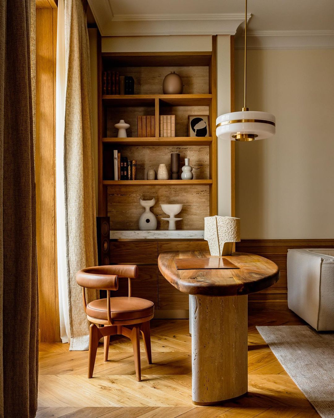 Interior of a home office with a wooden desk featuring a travertine base, a leather chair, and a built-in bookshelf with decorative objects.