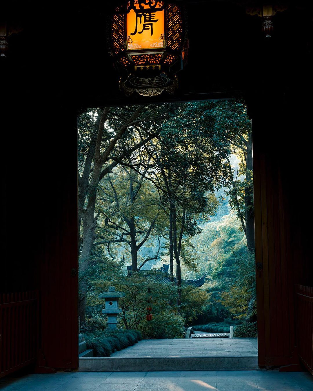 View through doorway with hanging lantern in the foreground to greenery and a traditional Asian building in the distance.