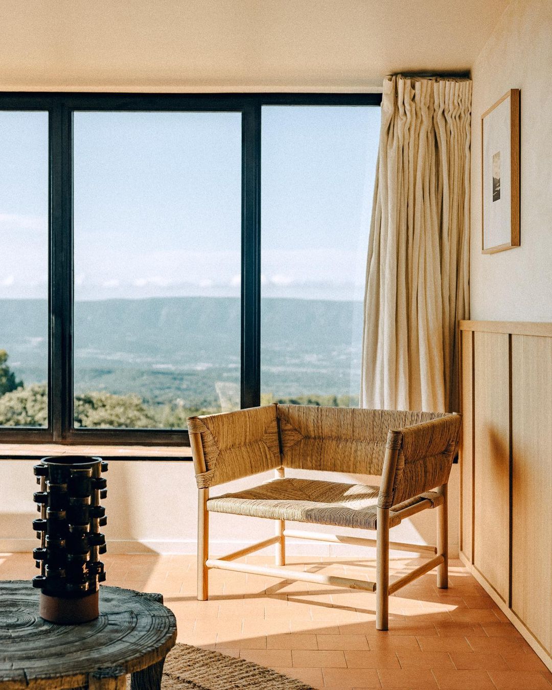 Interior with a woven armchair, round table, and scenic view through a large window, showcasing a blend of textures and natural light.