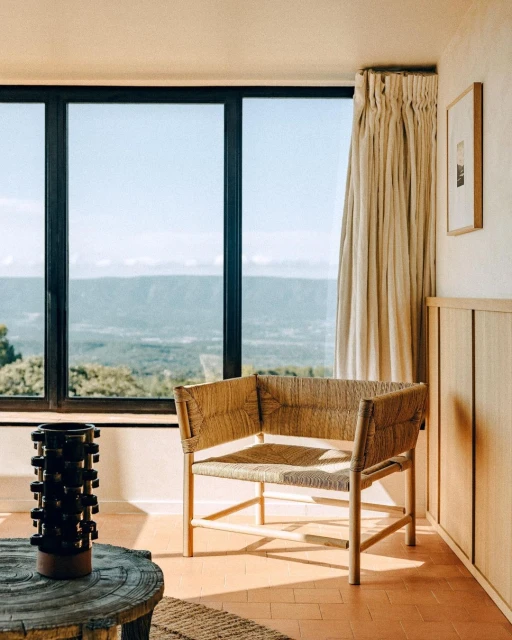 Interior with a woven armchair, round table, and scenic view through a large window, showcasing a blend of textures and natural light.