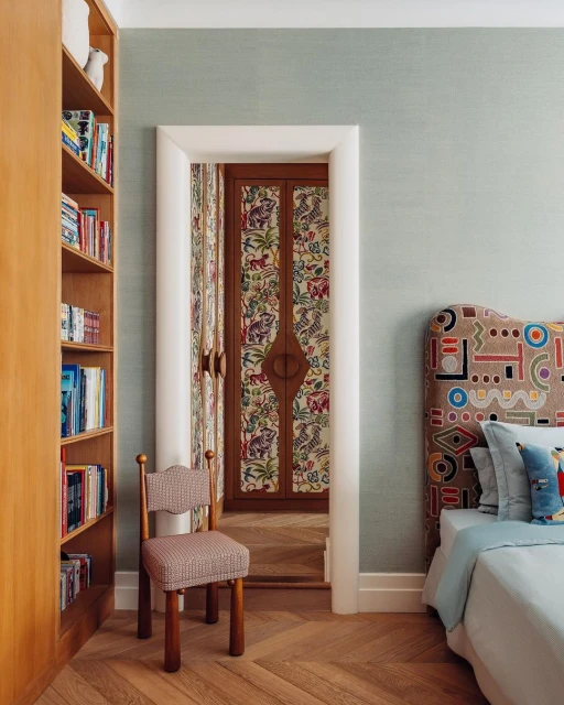 Interior bedroom view with a bookshelf, doorway with patterned double doors, chair, bed with an elaborate headboard, and parquet floor.