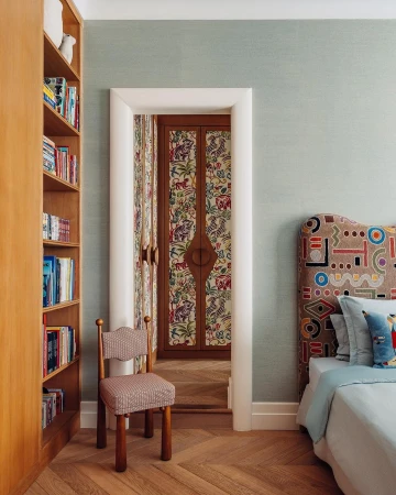 Interior bedroom view with a bookshelf, doorway with patterned double doors, chair, bed with an elaborate headboard, and parquet floor.