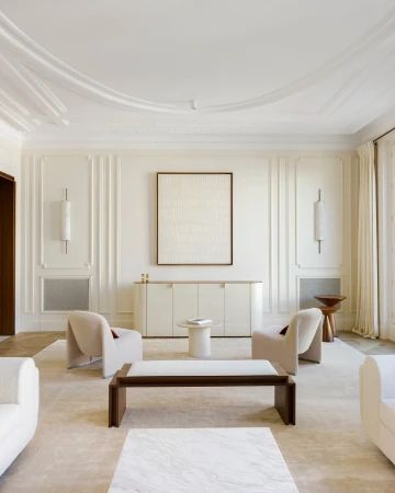 Bright and neutral living room with upholstered chairs, a marble coffee table, and an abstract art piece hanging on the wall.