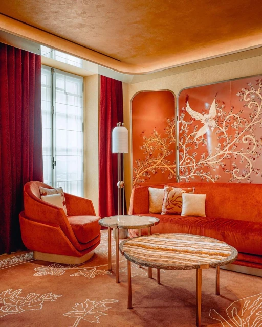 Elegant living room interior featuring an orange velvet sofa, armchair, round tables, patterned carpet, and decorative wall panel.