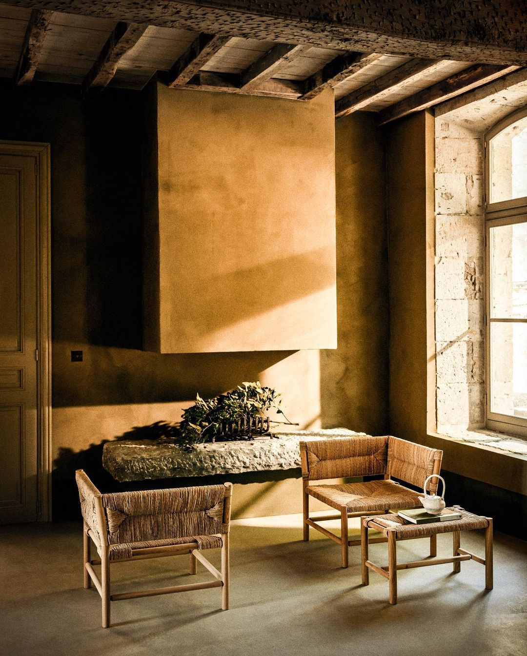 Interior design with straw chairs and stone table, featuring a rustic fireplace, set against textured walls and natural light from a window.