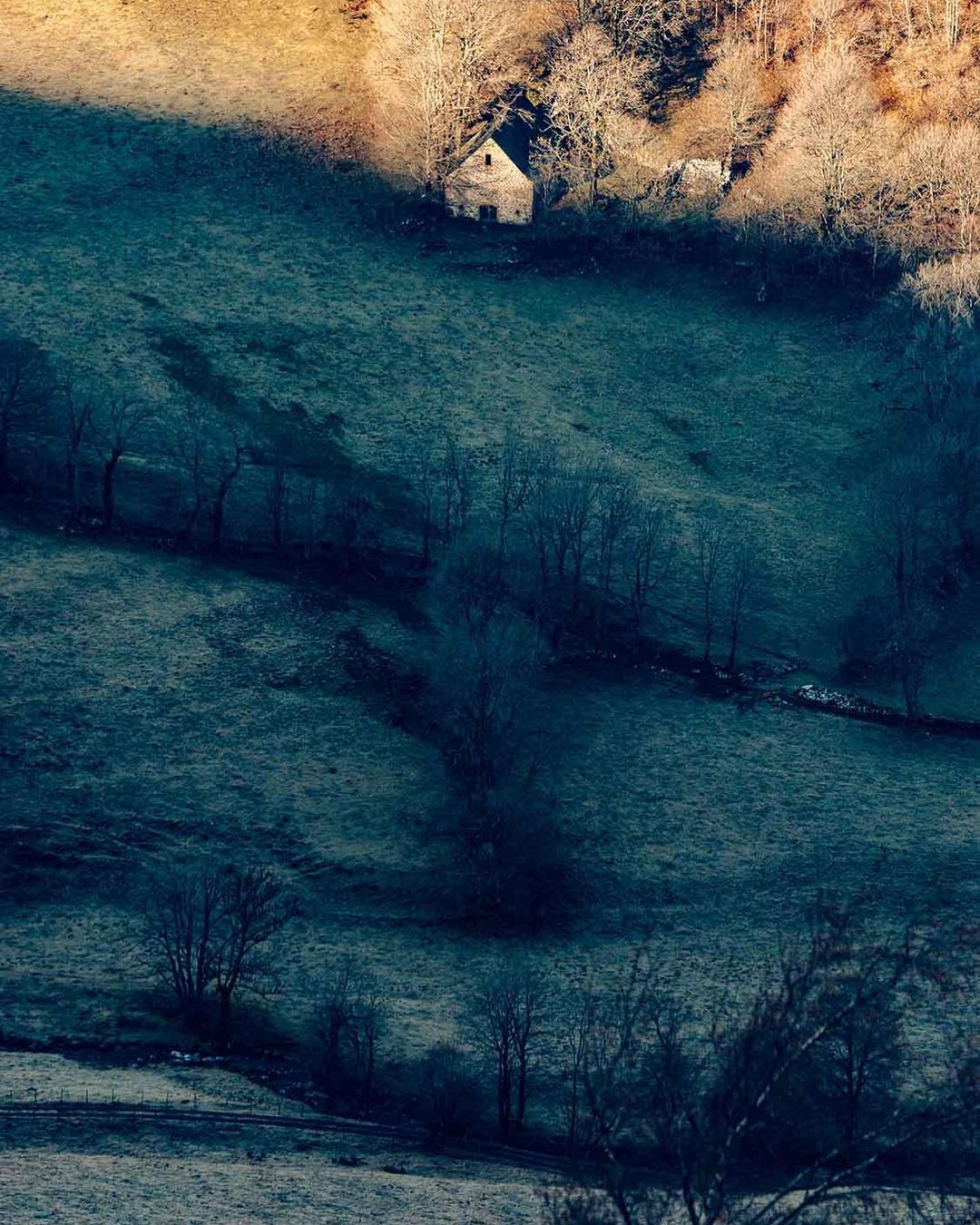 Distant stone house in a valley surrounded by frost-covered fields and leafless trees on a cold morning with the sun highlighting the peak.