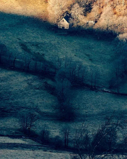 Distant stone house in a valley surrounded by frost-covered fields and leafless trees on a cold morning with the sun highlighting the peak.