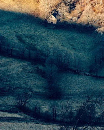 Distant stone house in a valley surrounded by frost-covered fields and leafless trees on a cold morning with the sun highlighting the peak.