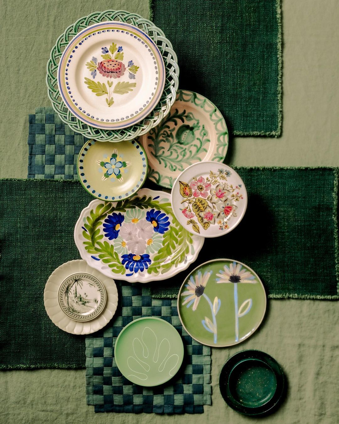 Overhead shot showcases an arrangement of patterned ceramic plates on a patchwork of green fabric squares, creating a textured, artistic composition.