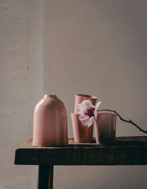 Still life arrangement featuring a pale pink vase, several cups, and a single white orchid on a rustic wooden surface against a neutral background.