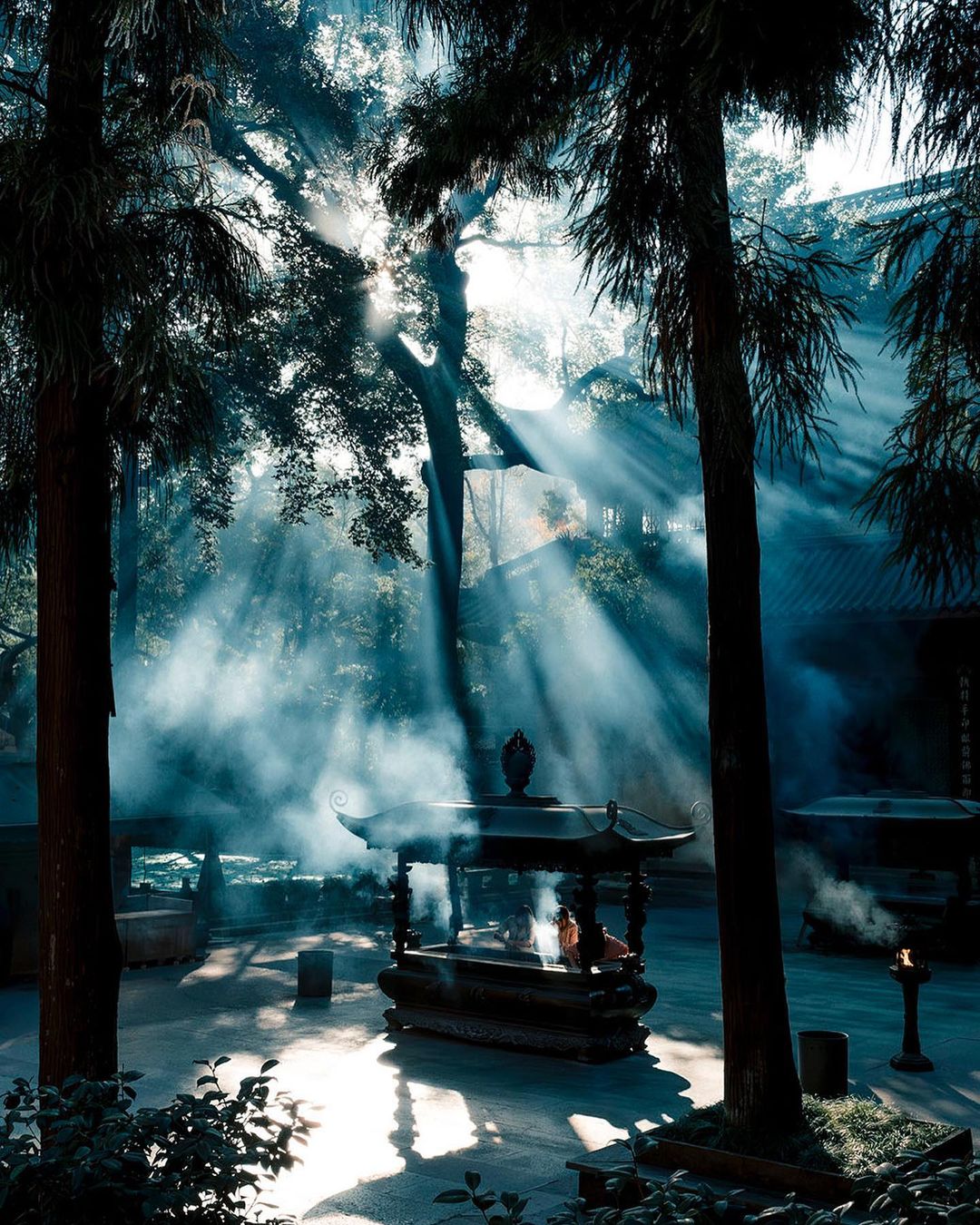 Sunlight streams through trees onto an ornate incense burner at a temple. Mist and fog add a serene, spiritual atmosphere to the landscape.