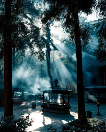 Sunlight streams through trees onto an ornate incense burner at a temple. Mist and fog add a serene, spiritual atmosphere to the landscape.