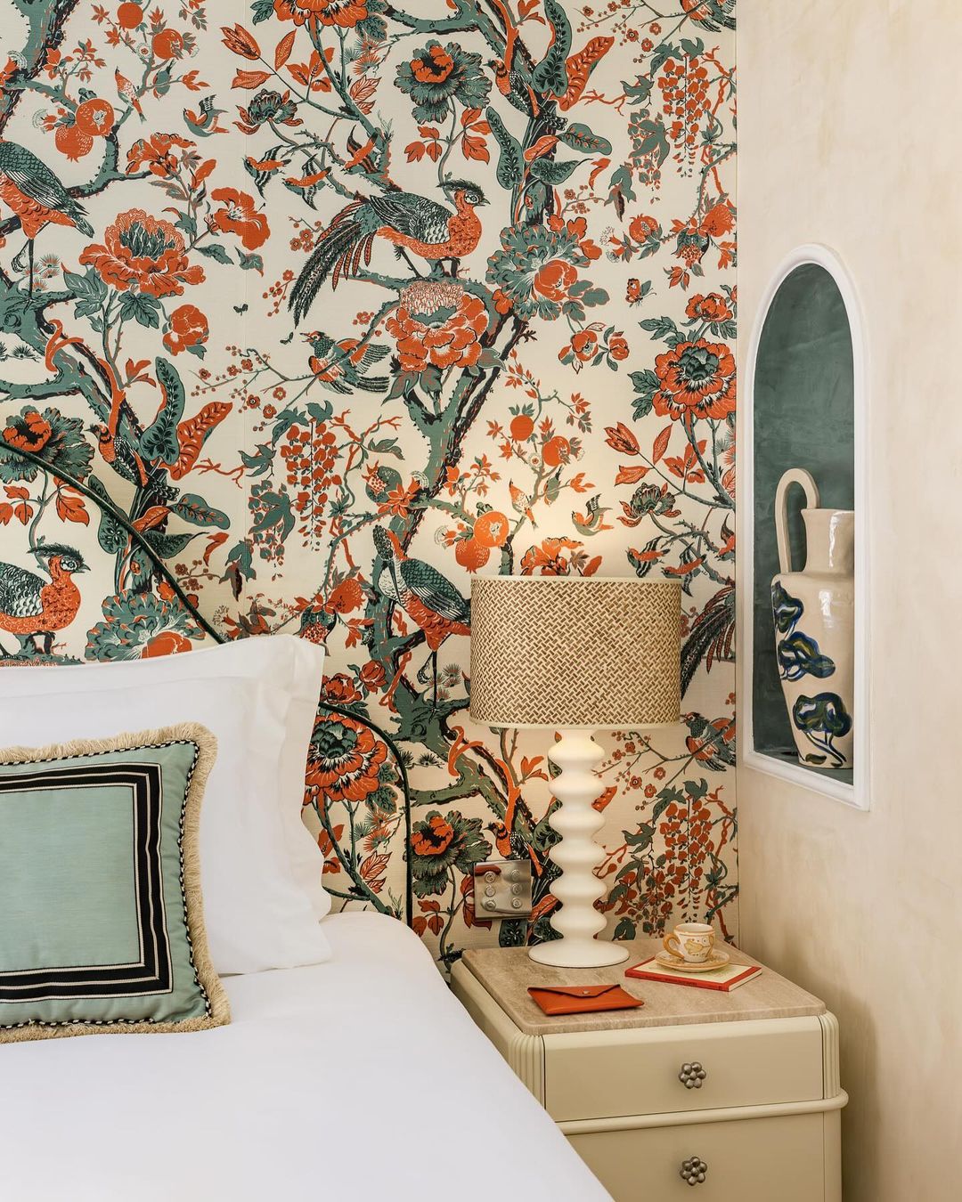 Bedroom interior with floral wallpaper, bedside table, lamp, and arched alcove featuring a white vase with blue floral design.