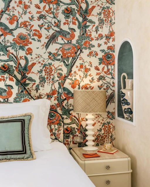 Bedroom interior with floral wallpaper, bedside table, lamp, and arched alcove featuring a white vase with blue floral design.