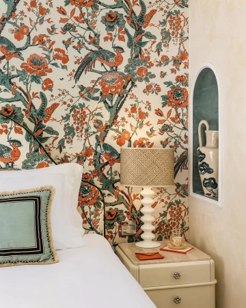 Bedroom interior with floral wallpaper, bedside table, lamp, and arched alcove featuring a white vase with blue floral design.