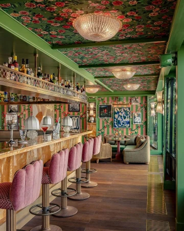 Interior of an ornate bar area, showcasing pink bar stools, a wooden bar, and decorative floral wallpaper and ceiling decor.
