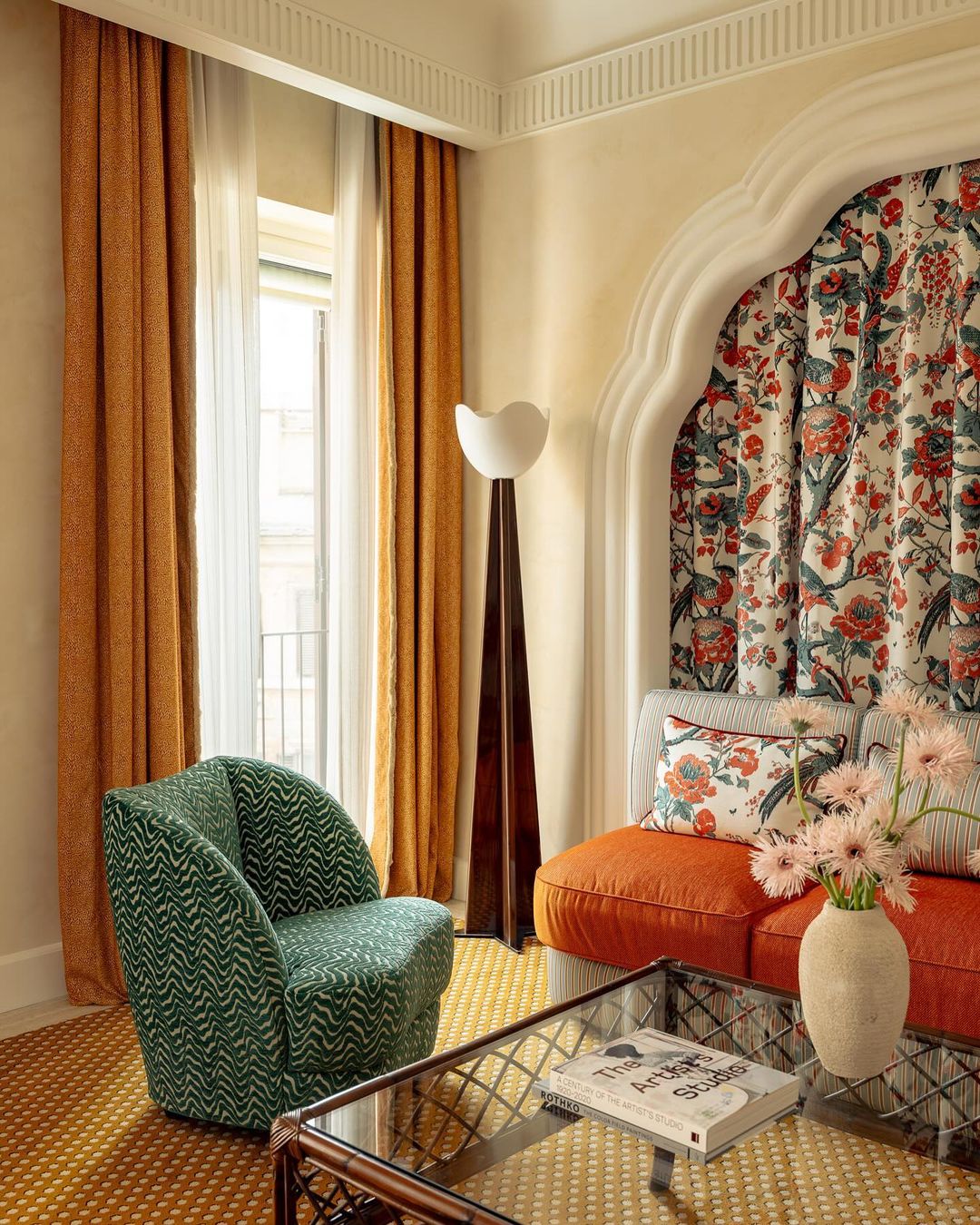 Living room interior with green armchair, orange sofa with floral fabric backdrop, floor lamp, glass coffee table, and patterned rug.