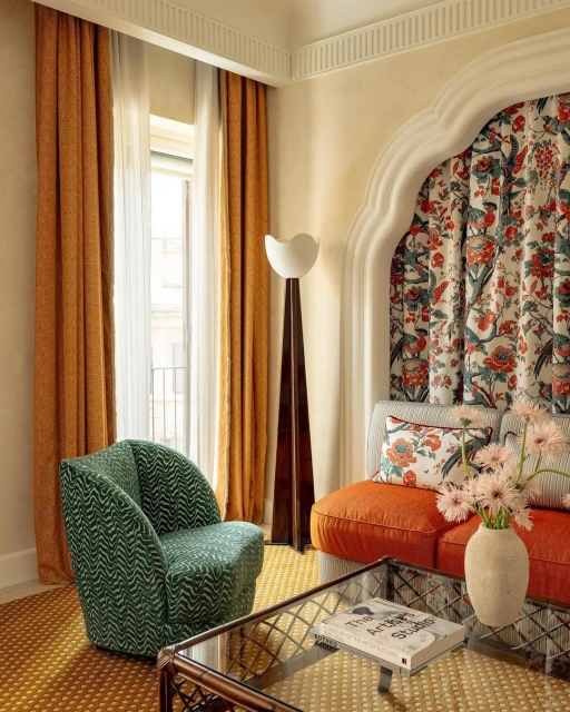 Living room interior with green armchair, orange sofa with floral fabric backdrop, floor lamp, glass coffee table, and patterned rug.