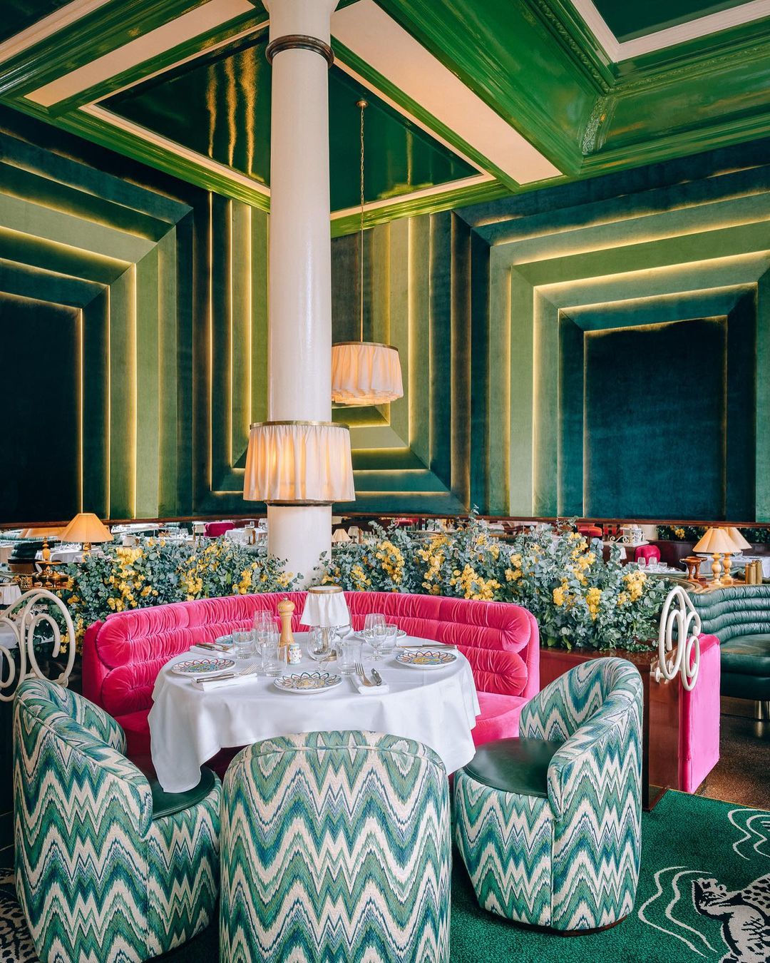 Elegant restaurant interior showcasing geometric green walls, white pillar, pink seating, patterned chairs, and a formally set round table.