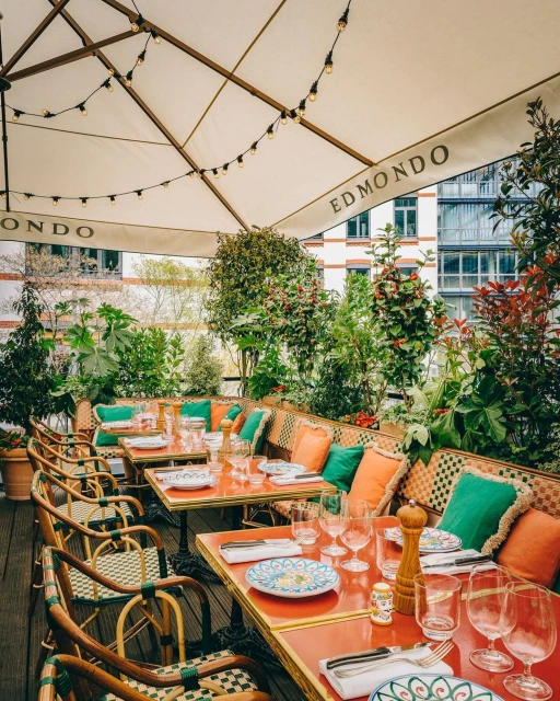 Outdoor restaurant seating under a white awning, tables set with plates and glassware, surrounded by plants and greenery.