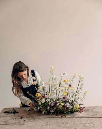 Adult female arranges a large, natural floral centerpiece with yellow, white, and red flowers on a rustic wooden table indoors.