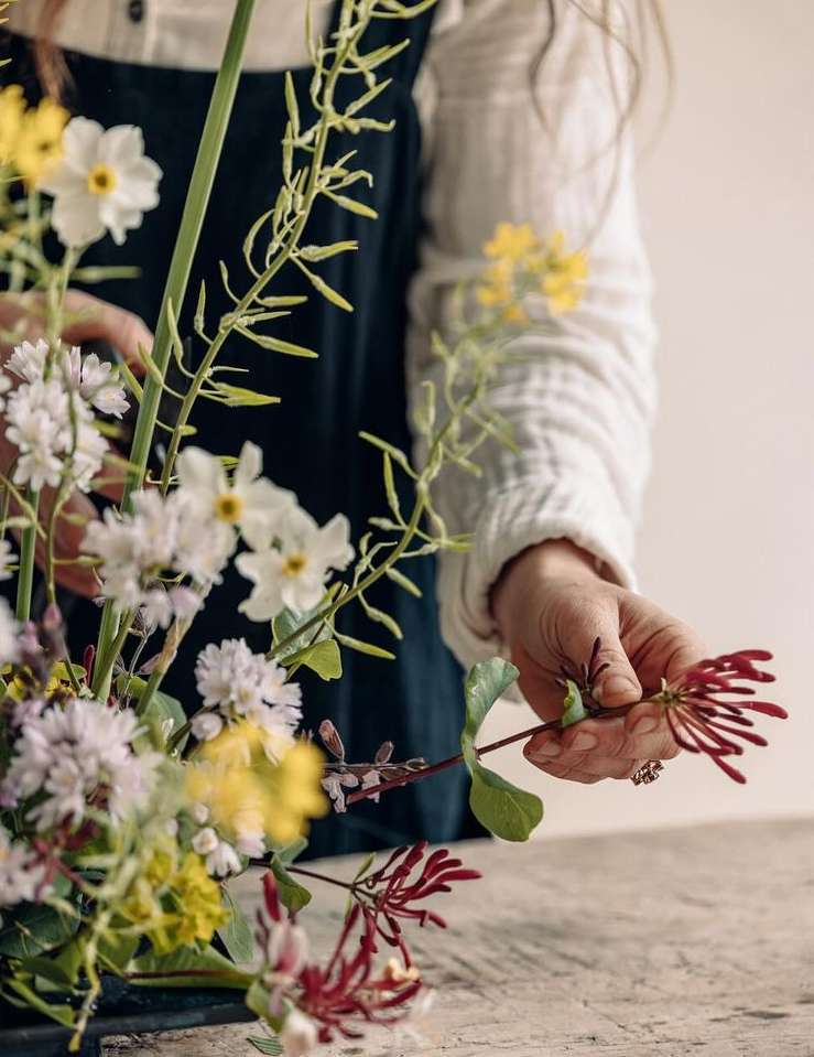 An artistic close-up features a woman's hands arranging a delicate floral bouquet with white, yellow, and red blooms on a neutral surface.
