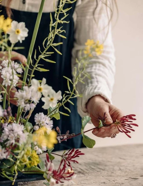 An artistic close-up features a woman's hands arranging a delicate floral bouquet with white, yellow, and red blooms on a neutral surface.